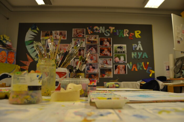 Ett kreativt aleljéutrymme med bord och material i förgrunden och en vägg fylld av färgglada konstverk och bokstäver i bakgrunden.