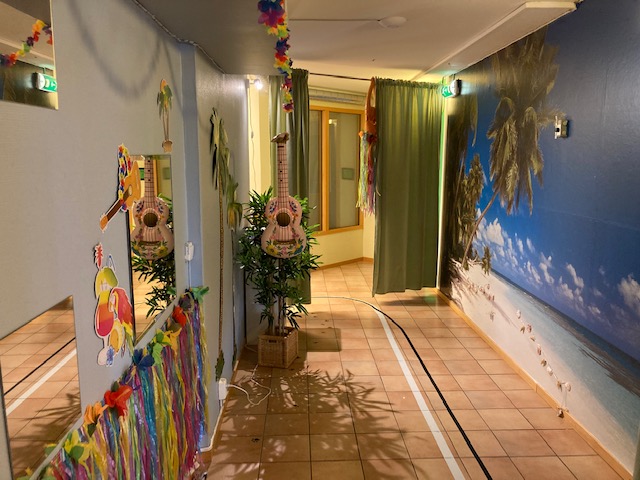 Korridor där ena väggen har en tapet som föreställer en vit strand, blått hav och några palmer. Andra väggen är hawaii-inspirerad med tygremsor i olika färger.