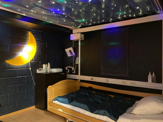 Säng i ett rum med mörkmålade väggar. En månlampa på ena väggen och små lampor i taket som en stjärnhimmel.