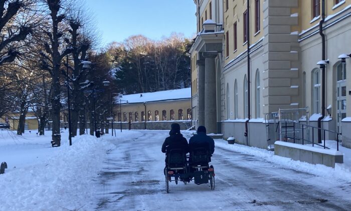 Snötäckt stadsmiljö med en byggnad längs ena sidan och träd längs den andra. Två personer färdas på en side-by-side cykel på den snöiga vägen.