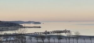 Utsikt över kust och hav i mjukt kvällsljus med piren i askimsviken som sträcker sig ut i vattnet. Träd och strandlinje syns i förgrunden och landkonturer i bakgrunden.
