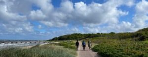 Kustlandskap med hav till vänster och grön natur till höger.En stig löper längs stranden och några personer promenerar under en himmel med spridda moln.