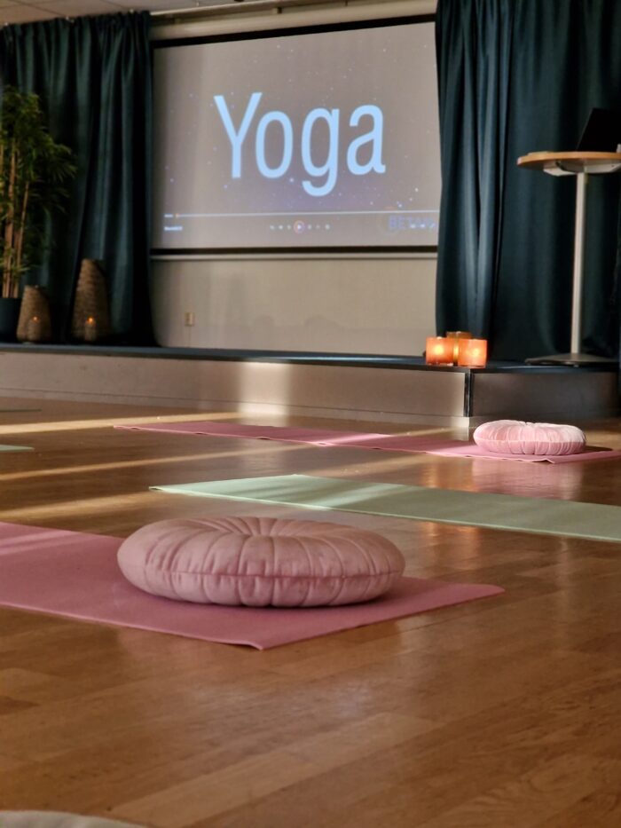 Sal med yogamattor och sittkuddar på golvet,scen och duk med texten yoga på väggen i bakgrunden.