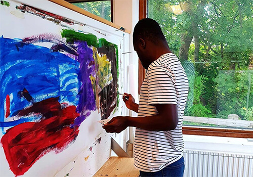 En person som målar en färgstark målning nära ett fönster med mycket grönska utanför.