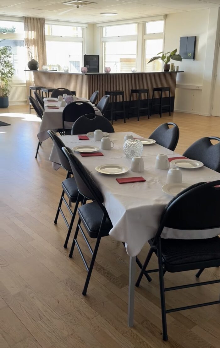 Långbord dukat med tallrikar och servetter samt svarta stolar runtom i ett ljust rum med fönster och serveringsyta i bakgrunden.