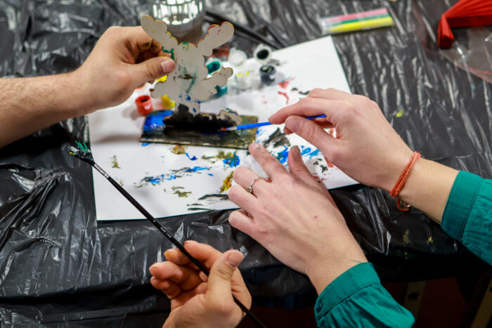 Flera händer målar och pysslar tillsammans vid ett bord täckt med skyddsplast.Penslar,färg och små dekorationer ligger framme runt ett pågående kreativt arbete.