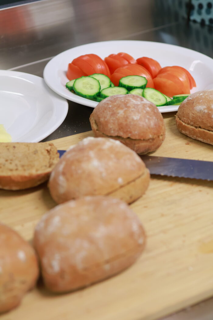 Frallor på skärbräda med kniv samt tallrik med skivade gurkor och tomater i bakgrunden.