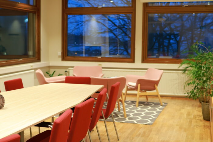 Ljust konferensrum med flera bord och röda stolar, stora fönster mot utsidan och en grön krukväxt i hörnet.