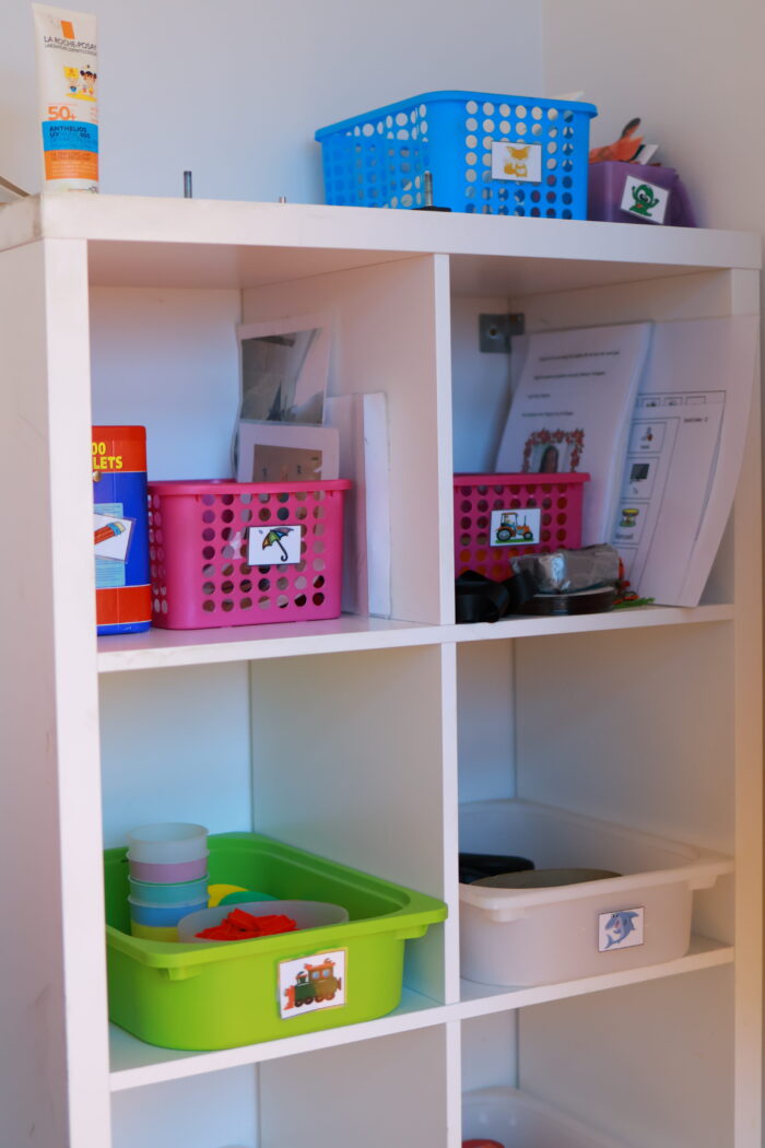 En vit hylla med flera fack som innehåller färgglada plastlådor och korgar med material och pärmar. Lådorna är märkta med bilder för sortering och struktur.