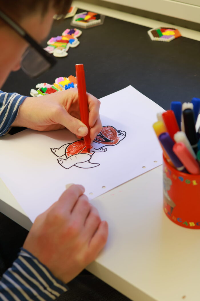 Närbild av en person som färglägger en teckning med röd penna vid ett bord,med dler färgpennor och pysselmaterial bredvid.