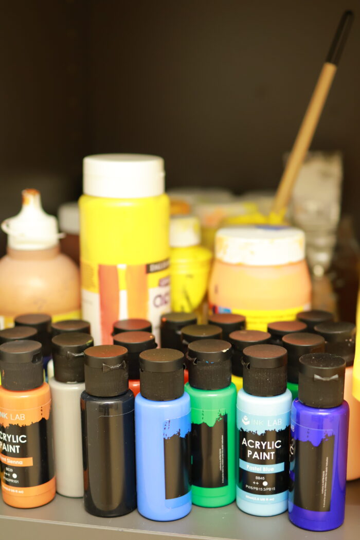 Flera flaskor med hobbyfärg och målarfärg i lika kulörer samt burkar och penslar på ett bord.