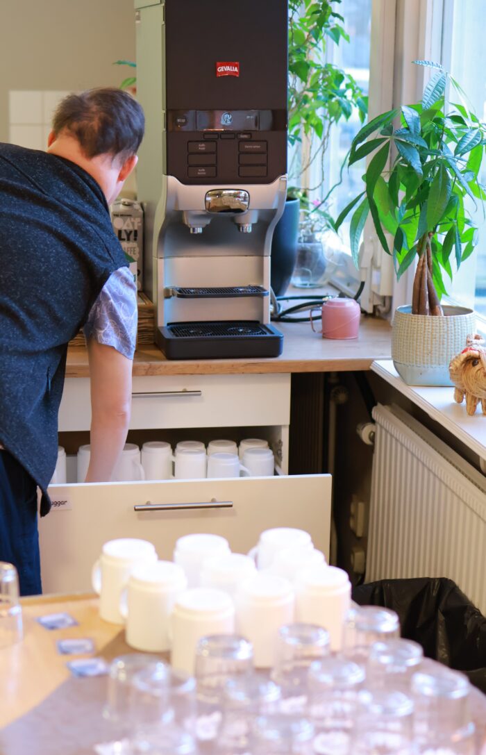 Person arbetar vid köksbänk med kaffemaskin i bakgrunden och glas samt muggar uppställda på bordet i förgrunden,växt vid fönster bredvid.