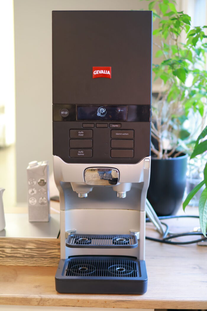 Kaffemaskin placerad på köksbänk med muggar bredvid och en grön krukväxt vid fönstret bakom.