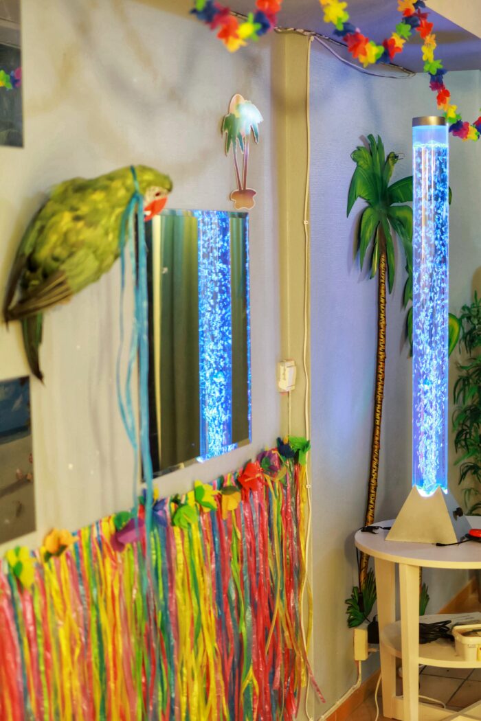 Hawaii-inspirerad vägg i en korridor för sinnesstimulans bestående av färgglada dekorationer och material. En papegoja i förgrunden och ett ljusrör med fiskar och bubblor.