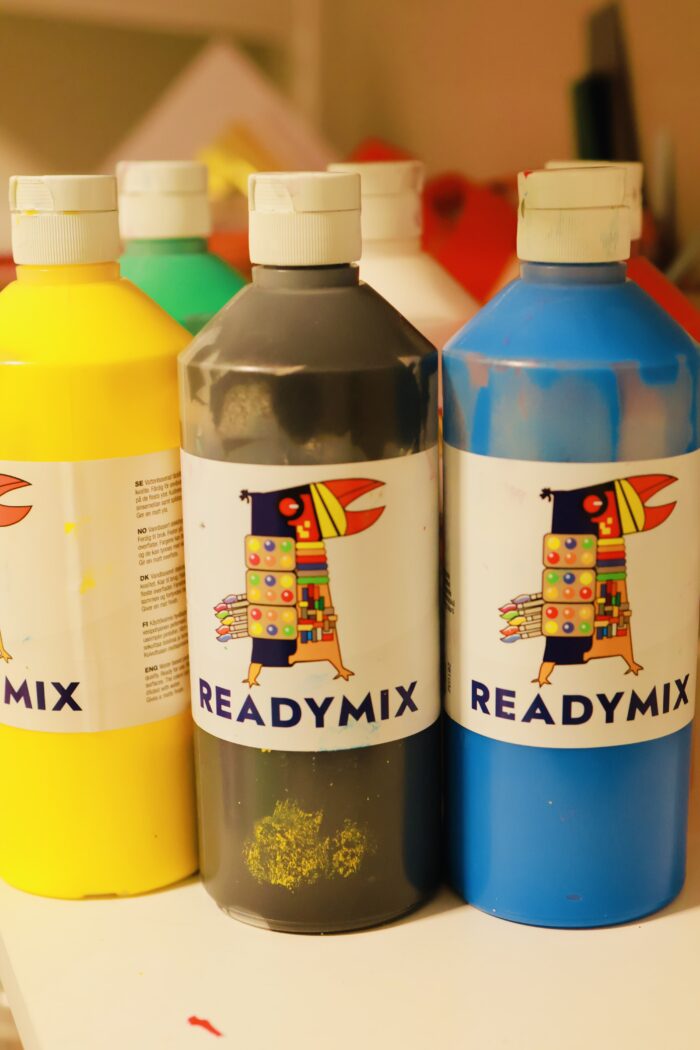 Närbild av tre flaskor med hobbyfärg i olika färger som står på ett bord med färgglada klossar suddigt i bakgrunden.