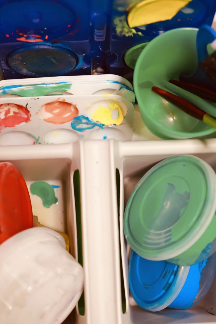 Hylla med plastlådor och burkar med lock i olika färger,fyllda med material och leksaker. Förvaring i ett aktivitetsrum.