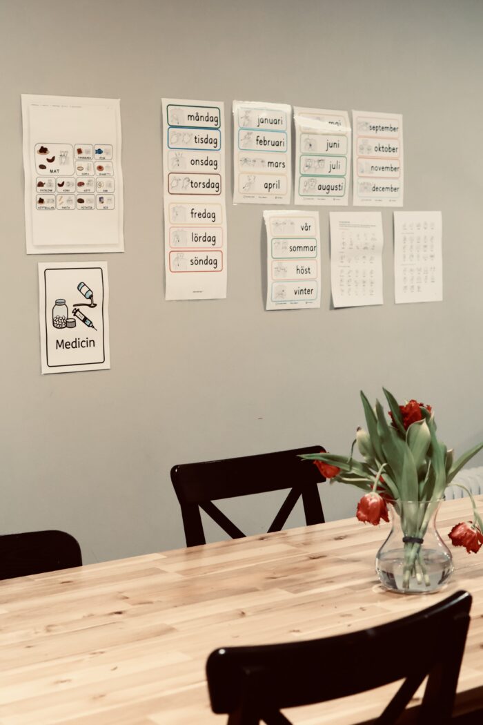 Ett aktivitetsrum med träbord och stolar. På väggen sitter flera uppsatta scheman eller informationsblad och på bordet står en vas med blommor.