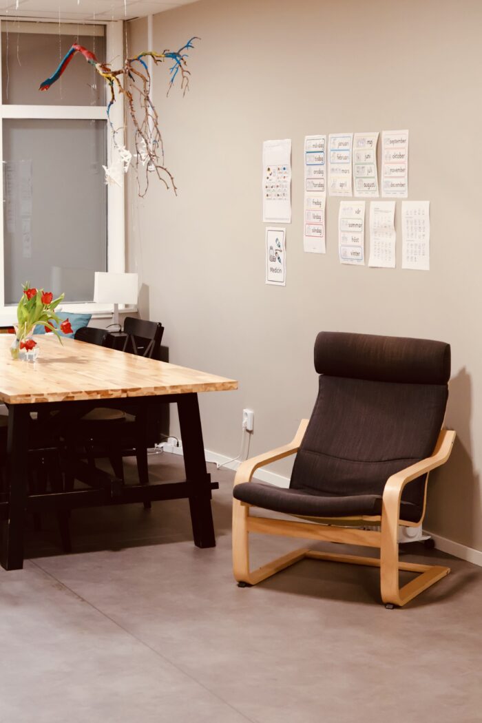 Ett lugnt rumshörn med ett bord och material till vänster och en bekväm fåtölj till höger. På väggen sitter bildstöd, vilket ger intryck av ett arbets eller samtalsrum.