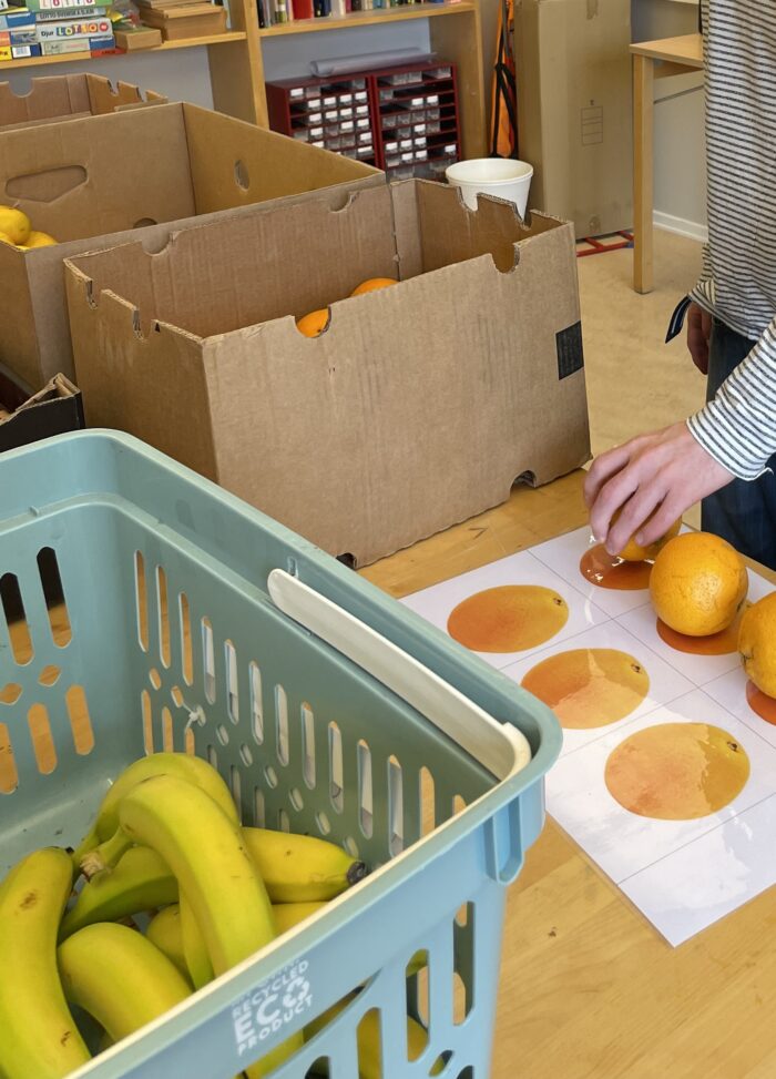 Korg med bananer på ett bord och en person som sorterar apelsiner på målade apelsiner, med kartonger och hyllor i bakgrunden.