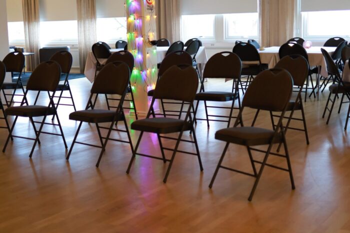Sal med flera uppställda svarta stolar i rader på trägolv, men fönster längs väggen och en färgglad ljusdekoration i mitten.
