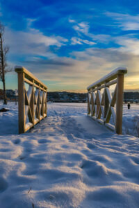 Snötäckt mark med en träbrygga som leder ner mot vatten. Himlen är blå med moln och kvällsljus i horisonten.