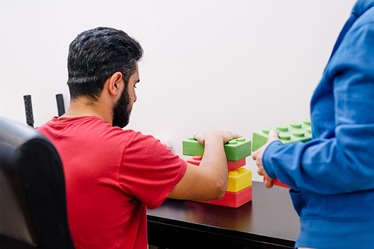 En person sitter vid ett bord och arbetar med färgglada klossari rött, gult och grönt. En annan person står bredvid och ger stöd. Inomhusmiljö med fokus på en praktisk aktivitet.