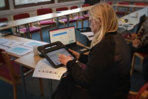 En person sitter vid ett arbetsbord och använder en samtalsplatta. Det finns även dator och flera olika dokument på bordet