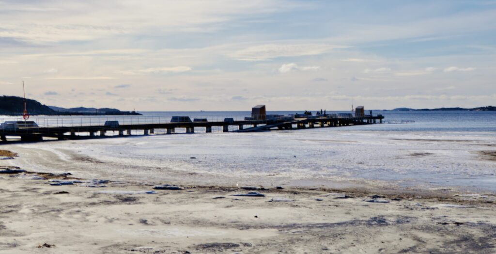 Sandstrand med den långa piren som sträcker sig ut mot Askimsviken under en ljusblå himmel