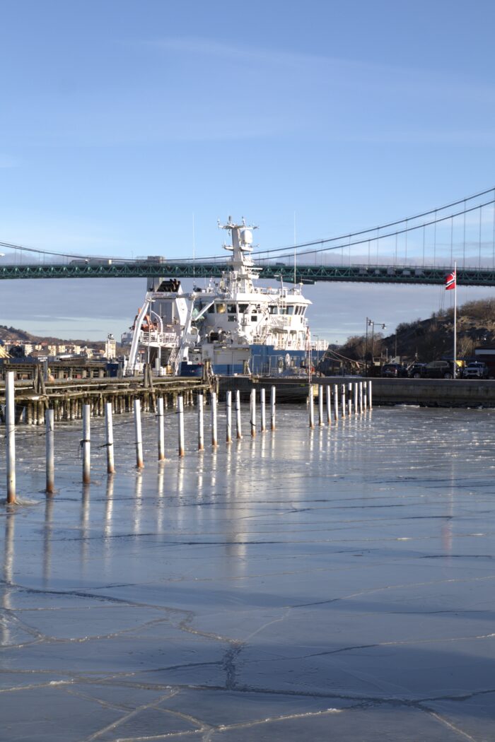 Vid bryggkanten. Pålar i vattnet i förgrunden och Älvsborgsbron och en båt i bakgrunden. Blå himmel och isflak i älven
