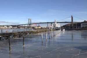 Vy över vatten med bryggor och stolpar i förgrunden samt Älvsborgsbron som sträcker sig över vattnet i bakgrunden.Stadsmiljö vid hamn under klarblå himmel.