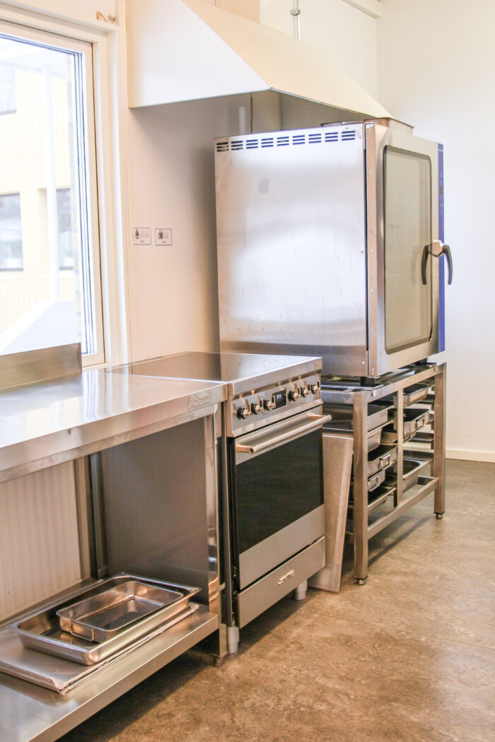 Stor varmluftsugn på en ställning för plåtar under. Till vänster om ugnen står en spis och sedan en arbetsbänk. Allt i rostfritt