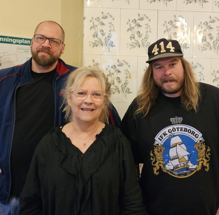 Tre personer poserar inomhus framför en vägg med ljusa kakelplattor med blommönster. En kvinna står i mitten och ler mot kameran. Bakom henne står två män – en till vänster med glasögon, kort hår och skägg i mörk jacka, och en till höger med långt hår, mustasch och skägg, svart keps med siffrorna “414” och en svart tröja med ett skeppsmotiv